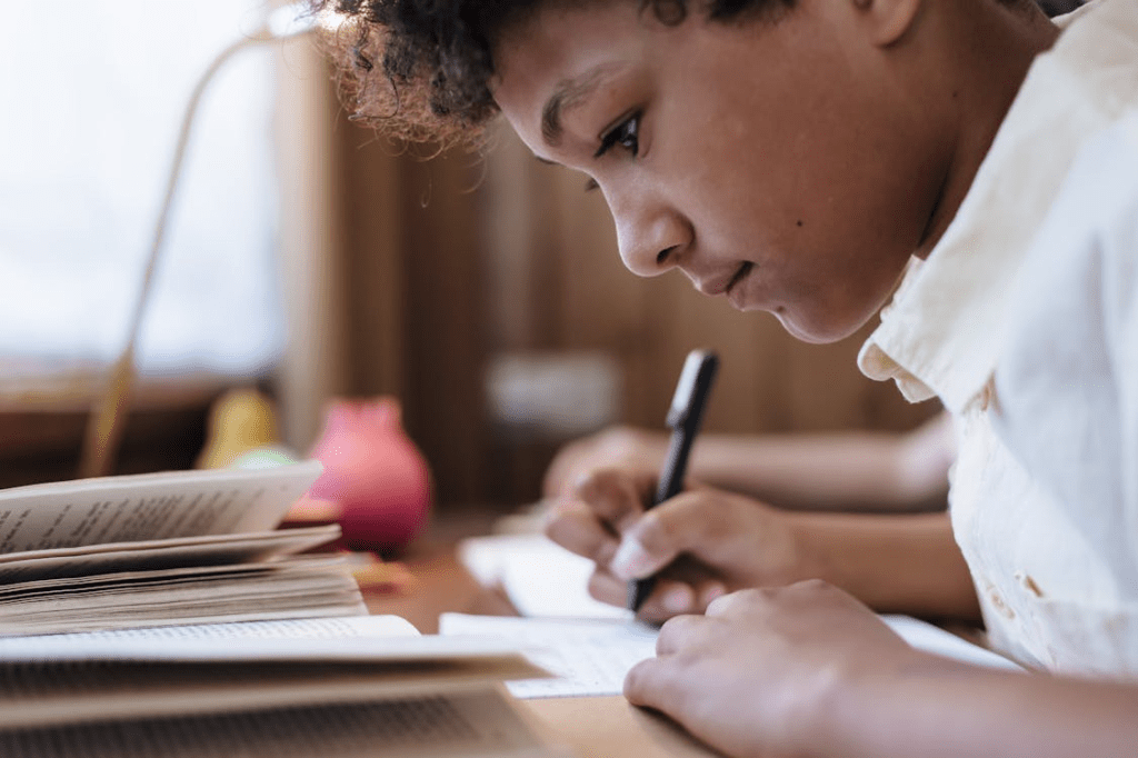 Un niño concentrado escribiendo en una hoja, rodeado de libros en una mesa de estudio.